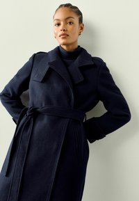 Manteau en laine navy à ceinture, avec un col large et des poches latérales. Tissu texturé avec un léger brillant.