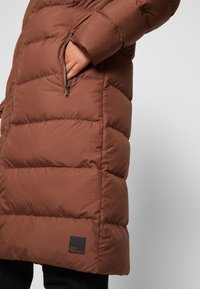 Brun puffjacka med sydda sektioner, sidoficka med dragkedja och logotag. Slät textur och medellång design, lämplig för kallt väder.