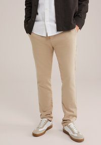 Beigu kokvilnas chinos ar šauru piegriezumu, plakanu priekšpusi un aizmugurējām kabatām, apvienoti ar baltām kedām ar brūnu zoli un pelēkiem akcentiem.