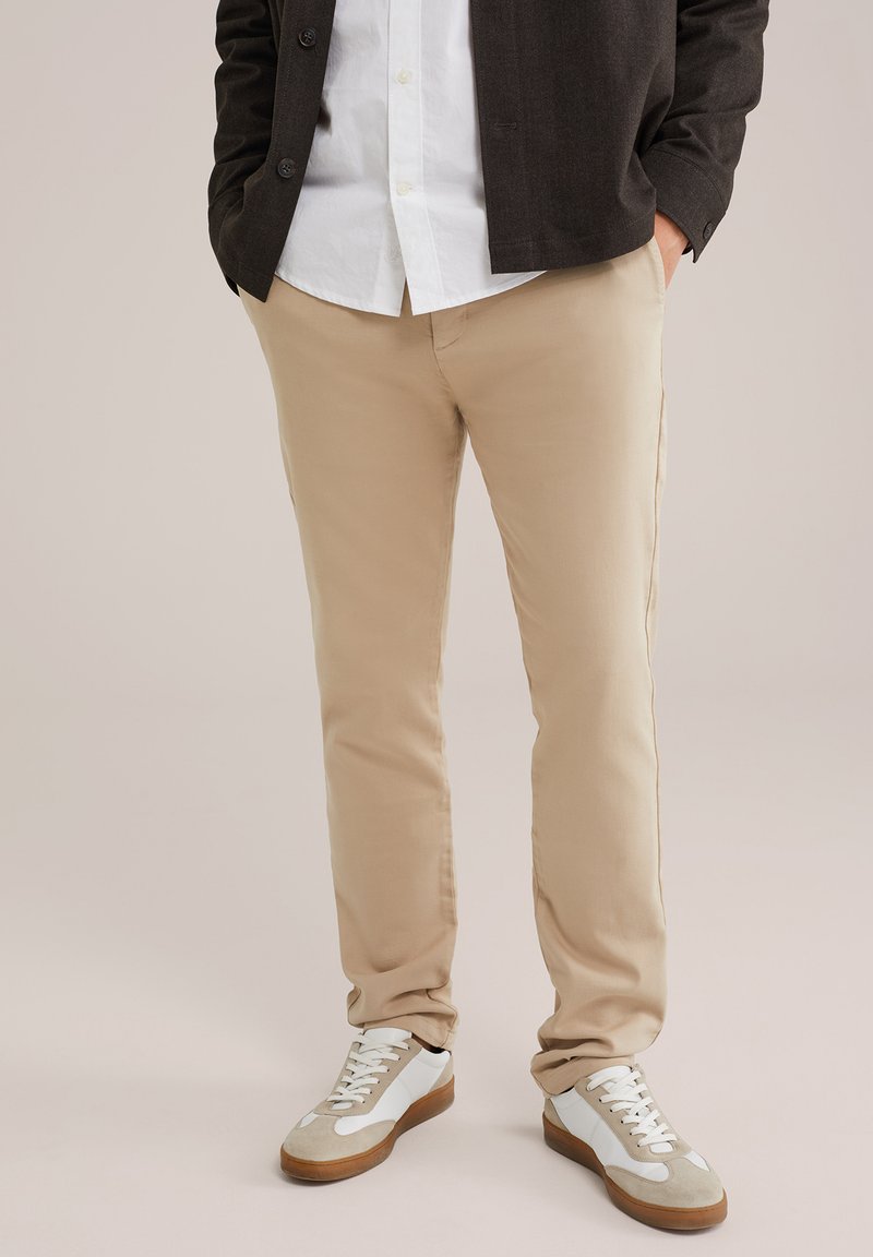 Beigu kokvilnas chinos ar šauru piegriezumu, plakanu priekšpusi un aizmugurējām kabatām, apvienoti ar baltām kedām ar brūnu zoli un pelēkiem akcentiem.