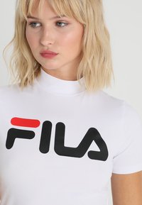 T-shirt blanc à manches courtes avec un col haut, arborant le logo FILA en noir et rouge. Texture lisse avec une coupe ajustée.