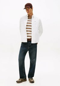Young man wearing a brown cap, white jacket over brown striped sweater, blue jeans, and brown sneakers, standing against a white background.