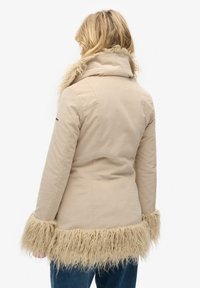 Beige jas met een fluffy, lichtbruine nepbontafwerking aan de manchetten en onderkant, met een hoge kraag en een getailleerde pasvorm.