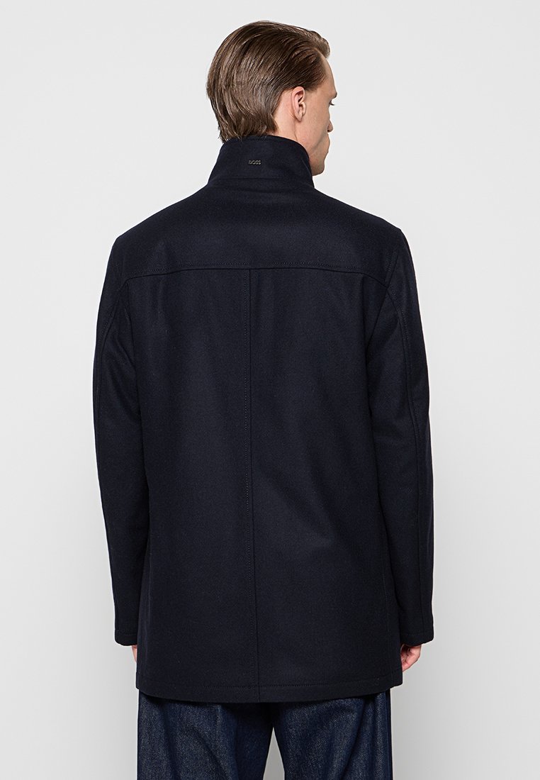 Cappotto in misto lana blu navy con colletto alto, vestibilità sartoriale e texture liscia, caratterizzato da cuciture minime e una silhouette pulita.