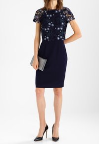Abito blu navy con corpino in pizzo floreale, maniche a sbuffo, gonna aderente e un tessuto testurizzato. Abbinato a una pochette argentata e tacchi neri.