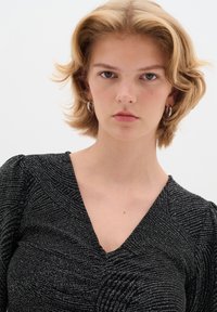 Top nero con scollo a V, tessuto texturizzato con un motivo argentato a vortice. Maniche a sbuffo e silhouette aderente. Orecchini a cerchio metallici.