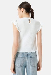 Top blanco texturizado con mangas acampanadas, espalda con abertura en forma de lágrima y dobladillo con puntas. Combinado con jeans de mezclilla azul claro con bolsillos traseros.