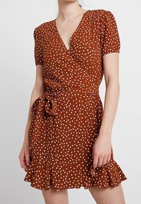 Robe enroulée marron avec des pois blancs, manches courtes bouffantes, une ceinture à la taille et un ourlet évasé. Fabriquée en tissu léger.