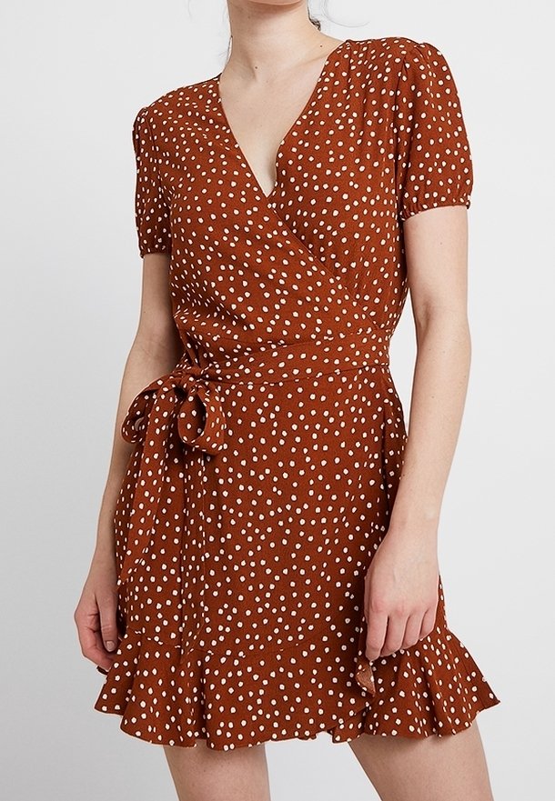 Robe enroulée marron avec des pois blancs, manches courtes bouffantes, une ceinture à la taille et un ourlet évasé. Fabriquée en tissu léger.