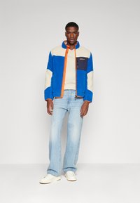 Blå fleecejakke med en beige brystplate og marineblå lomme. Har oransje glidelås og krage. Sammen med lyseblå jeans og hvite sko.