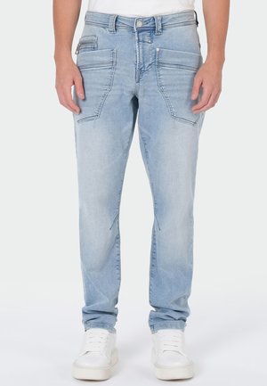 Person wearing light blue jeans with multiple front pockets and white sneakers, standing against a plain background.