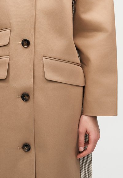 Μπεζ παλτό με μαύρα κουμπιά και δύο τσέπες με καπάκι, φορεμένο από άτομο που δείχνει το δεξί χέρι και βραχίονα πάνω σε ανοιχτό φόντο.