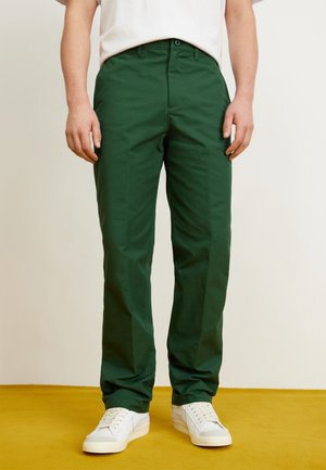 Personne portant un pantalon droit vert foncé et des baskets blanches debout sur un sol jaune contre un mur clair.