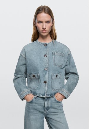 Mladá žena ve světle modré džínové bundě s velkými předními kapsami a odpovídajícími džínami, stojící s rukama v kapsách, neutrální pozadí.