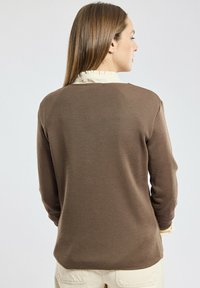 Maglione marrone lavorato a maglia con maniche lunghe, texture a coste e vestibilità morbida. Un colletto arricciato di colore crema è visibile sotto.