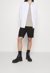 Bílá košile s knoflíky přes olivové pruhované tričko, černé kraťasy a černé Chelsea boty s žlutým akcentem na jednobarevném pozadí.