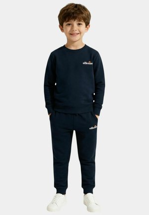 Jeune garçon portant un sweat-shirt ellesse bleu marine et un pantalon assorti, avec des baskets blanches, debout les mains dans les poches, souriant.
