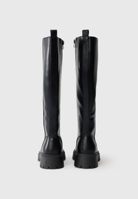 Cizme din piele neagră, până la genunchi, cu o textură netedă, dotate cu un fermoar pe spate și talpă din cauciuc masivă pentru stabilitate.