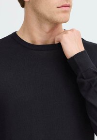 Fekete kötött pulóver, kerek nyakkivágással, bordás mandzsettákkal és puha textúrával, a nyak és a felkar területének közeli képe.
