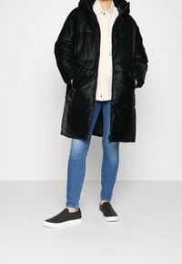 Cappotto imbottito nero con cappuccio, dotato di chiusura con zip e due tasche frontali, abbinato a jeans skinny blu e scarpe nere senza lacci.