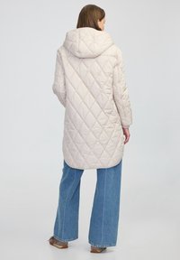 Padded béžový kabát s velkou kapucí, s prostorem na zádech, rukávy do lokte a střední délku ke stehnům. Spárován s kalhotami širokého střihu.