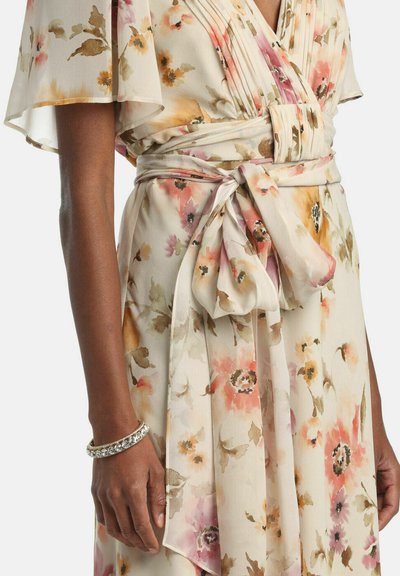 Közeli kép egy virágmintás, fodros ujjú és megkötős derékszíjas ruháról, sötét bőrű személyen, aki ezüst karkötőt visel.