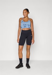 Top desportivo azul tie-dye com alças finas, combinado com calções de ciclismo pretos com textura em canelado. Sapatos de desporto pretos e meias completam o visual.