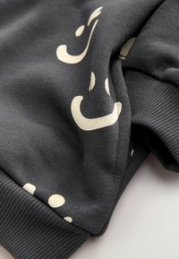 Tissu gris foncé avec des formes courbes abstraites et des points en blanc cassé, présentant des poignets côtelés et un détail de couture replié.