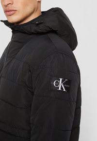 Svart puffjacka med huva, med kviltade sektioner och en framträdande Calvin Klein Jeans-logotyp på vänster ärm.