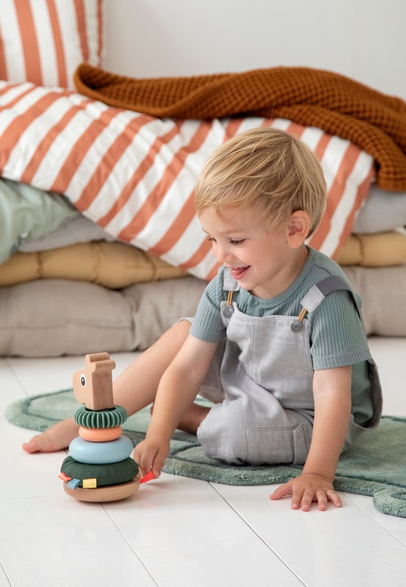 Tout-petit aux cheveux blonds assis sur un tapis vert, jouant et souriant avec un jouet empilable en bois et tissu sur un sol blanc.