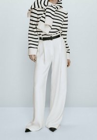Paski sweter w kolorze kremowym i czarnym, związany na szyi; białe spodnie o szerokich nogawkach z zakładkami; czarne buty na wysokim obcasie; minimalistyczny design.