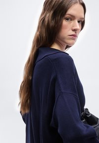 Νεαρή γυναίκα με μακριά καστανά μαλλιά που φοράει σκούρο μπλε πουλόβερ, κρατώντας μια μαύρη δερμάτινη τσάντα, κοιτάζοντας πάνω από τον ώμο της με φόντο λευκό.