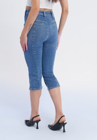 Denim capri džinsi ar pieguļošu dizainu, vidējiem vidukļa izmēriem un aizmugurējām kabatām, ar aplokām ap apakšu. Kombinēti ar melniem atvērtām kurpēm ar papēdi.