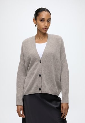 Vrouw draagt een beige gebreid vest met knopen over een wit topje en een zwarte rok, staand tegen een effen witte achtergrond.