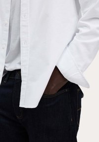 Weißes Hemd mit langen Ärmeln, das ein sauberes Design aufweist, in dunkle blaue Jeans mit sichtbaren Nähten gesteckt.