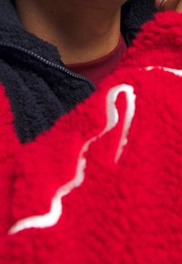 Červená fleecová bunda s texturovaným povrchem, s tmavě modrým límcem a bílým designovým akcentem, který diagonálně prochází látkou.