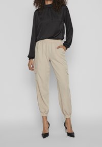 Blusa nera in satin con scollatura arricciata, abbinata a pantaloni cargo beige a gamba affusolata con tasche laterali e polsini elasticizzati, indossata con tacchi neri.