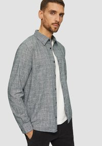 Grijze langesleeve geweven shirt met een gestructureerd ruitpatroon, knoopsluiting aan de voorkant en een borstzak. Gedragen over een wit T-shirt.