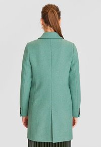 Cappotto verde in lana con colletto a scollatura, maniche lunghe e spacco posteriore. Presenta polsini con bottoni e una texture liscia. Vista posteriore mostrata.