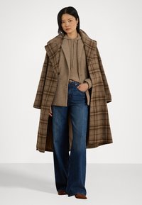 Bruine geruite wollen jas met een grote kraag over een beige gebreide trui en blauwe flared jeans. Heeft voorzakken en een ontspannen pasvorm.