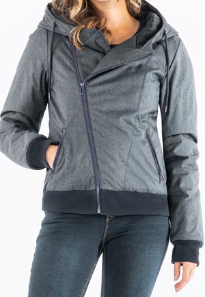 Kvinde iført en grå hættetrøje med asymmetrisk frontlynlås og lynlåslommer i siden, kombineret med mørkeblå jeans.