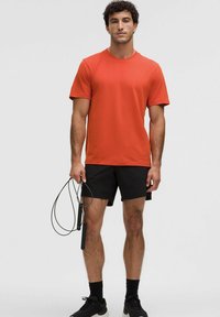 Tricou sportiv scurt, cu mânecă scurtă, de culoare roșie, fabricat dintr-un material ușor, asortat cu pantaloni scurți negri. Persoana ține o frânghie de sărit.