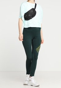 T-shirt cropped azzurro chiaro, leggings verde scuro con logo giallo, borsa in vita nera, sneakers bianche, abbigliamento sportivo casual.
