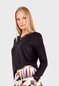Black long-sleeve top with a V-neckline, featuring a sheer trim. Paired with patterned, multicolored trousers showing geometric designs.