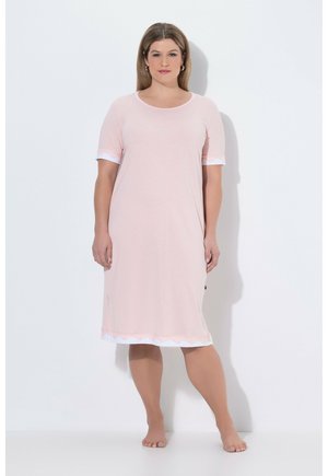 Mujer de pie descalza, vestida con un vestido rosa claro hasta la rodilla, con ribete blanco con borde ondulado en las mangas y el dobladillo, frente a una pared blanca.