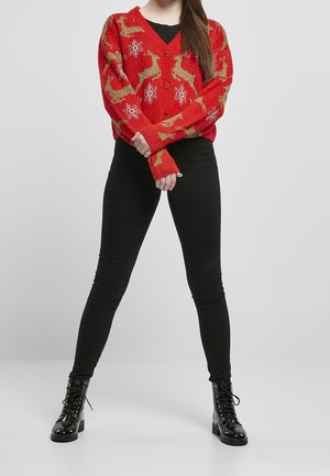 Kvinde iført rød julesweater med guld rådyr og hvide snefnug, sorte bukser og sorte snørestøvler, stående med armene over kors.