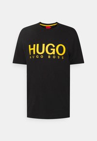 Camiseta negra de manga corta hecha de algodón. Presenta un logo grande en amarillo "HUGO" y un texto más pequeño "HUGO BOSS". Cuello redondo con ribete en contraste.