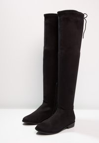 Bottes en suède noir hautes avec des talons bas, des orteils pointus, des fermetures éclair latérales et un détail de laçage à l'arrière, posées sur une surface blanche.