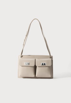 Sac à épaule beige avec deux poches avant, quincaillerie argentée, texture lisse et bandoulière réglable. Design épuré avec une esthétique minimaliste.