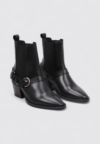 Bottines en cuir noir avec un bout pointu, un talon carré et des panneaux latéraux en élastique contrastant. Sangle décorative avec un détail de boucle ronde.